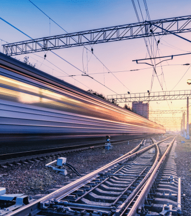 Train passant à grande vitesse sur des voies ferroviaires au crépuscule, avec caténaires et infrastructures électriques visibles